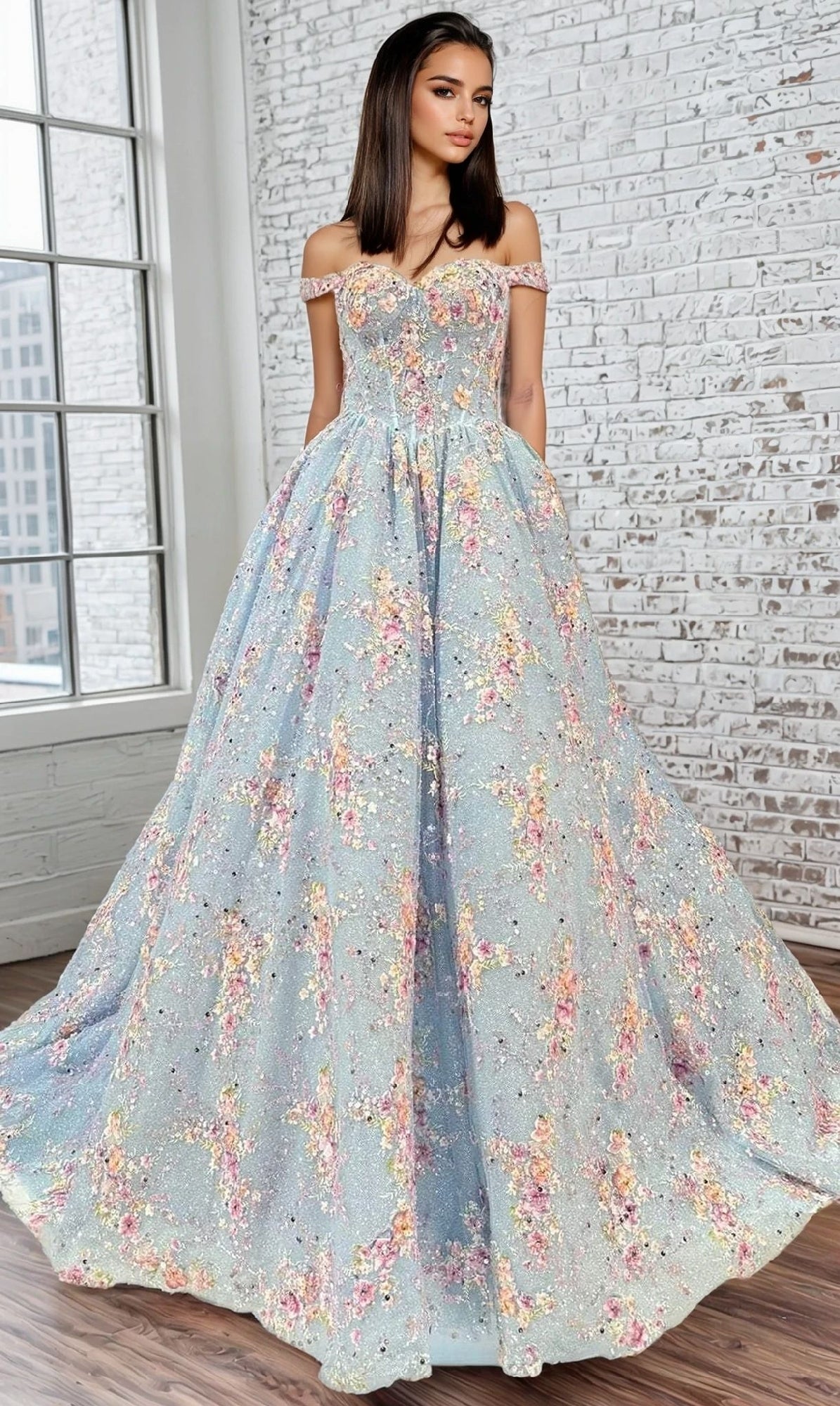 A woman poses indoors by large windows and a white brick wall, wearing the Portia and Scarlett PS25270 long prom dress—an off-the-shoulder gown with floral embellishments, a fitted corset bodice, and a full pastel-patterned skirt.